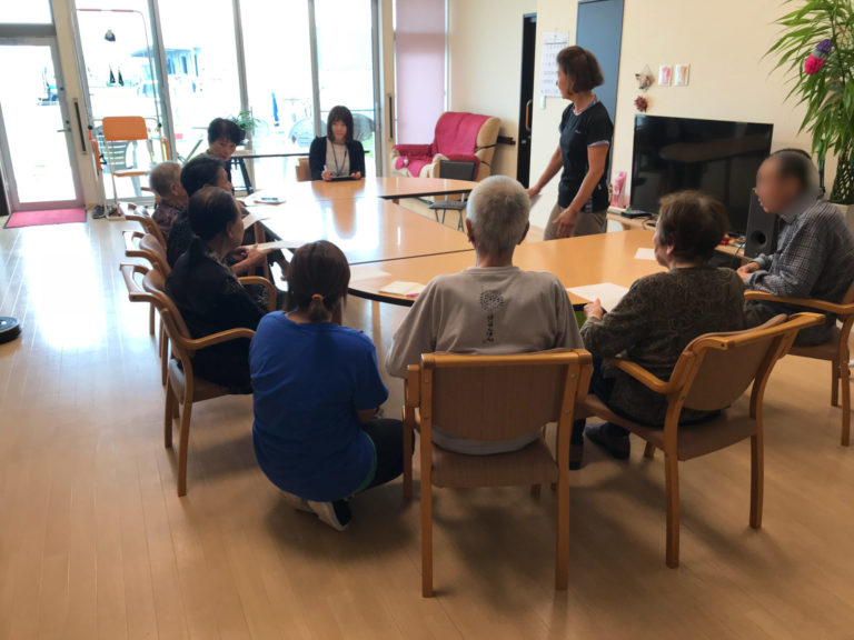 介護施設でのレクリエーション活動。入居者とスタッフが円形に座り、集団活動に参加している様子。